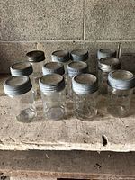Front view of twelve clear glass jars with zinc lids arranged in rows on rough wood surface against concrete block wall.