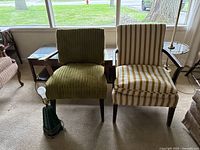 Photo showing green corduroy armchair and yellow and white striped armchair with retro green lamp in front on carpeted floor near window.