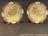 Two antique fruit bowls with hand painted floral design and gilded accents, purple rims with raised white and gold floral decorations, displayed side by side.