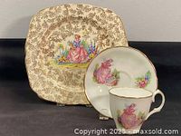 Photo showing the 1930s square Crinoline Lady plate with ornate floral background and pink crinoline lady image, alongside the 1950s teacup and saucer set with matching pink lady motif and gold trim.