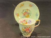 Front view of the pale green teacup with colorful hand painted flower design and gold trimmed handle, paired with matching saucer showing similar floral accents and scalloped gold rim.