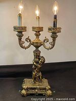 Front angled view showing ornate brass lamp with cherub holding up three light arms with candle-style bulb holders and decorative base.
