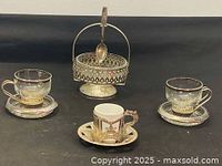 Photo showing two glass cups with silver handles and bases, one porcelain cup with silver holder, silver saucers, and decorative silver holder with spoon arranged on black surface.