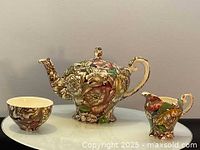Floral chintz teapot, creamer, sugar bowl and one cup shown on white surface against gray background.