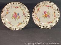 Pair of Schumann Bavaria antique porcelain plates with basket lace rims and hand painted floral designs on a white background, side by side on black surface.
