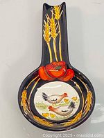 Full view of black ceramic spoon rest featuring hand painted golden wheat stalks on the handle and red poppy flower near the bowl rim, with a circular image of farm chickens and chicks inside the spoon bowl.