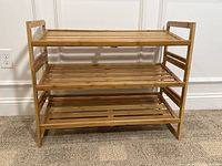 Photo of a three-tier bamboo shoe shelf with natural wood finish placed on carpeted floor near a white wall and electrical outlet.