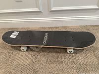 Side view of skateboard showing black grip tape with Hoodo branding on top deck and white wheels and black trucks on carpeted floor.
