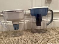 Two water pitchers placed on carpet floor, one transparent with white top and blue filter (Brita), the other transparent with white top and gray handle (Pur).