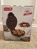 Front of boxed Dash Express Mini Donut Maker showing the open unit with 7 mini donuts cooking and a cooked donut assortment on a plate behind.