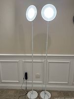 Front view of the white floor lamp with two circular LED lights glowing, showing the full height and design against a beige wall with white wainscoting.