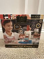 Front of FAO Schwarz Cake Me Away 15-piece cake pop maker set box showing child playing with cake pop maker and assembled cake pops on sticks