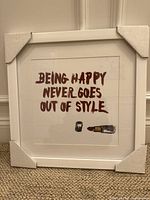 Front view of the framed art print leaning against a wall, showing the text and lipstick illustration clearly. Protective corners are on the frame edges.