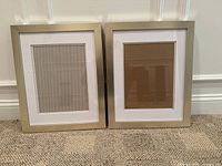 Two gold wooden picture frames standing on carpeted floor, each with white matting inside sized for 8x10 photos, showing reflections on glass surfaces.