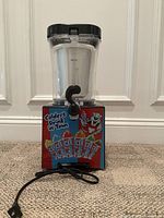 Front view of the ICEE Slushie Machine, showing multicolor branded base, transparent container with black lid, and power cord on a carpeted surface.