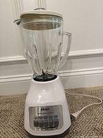 Oster Duralast Classic blender on carpeted floor. White base with several push buttons to control speed. Clear glass jar with lid and handle attached.