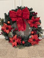 Front view of the 18 inch artificial green pine needle wreath decorated with red velvet bow, red poinsettia flowers, red berries, and red ornaments.