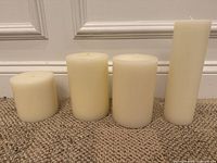 Four ivory pillar candles of different heights and diameters placed on carpet in front of a white wall baseboard, showing their sizes and shapes.