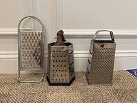Photo showing three stainless steel graters: one flat grater and two box graters with different sizes and handles, placed on carpet against a white wall.
