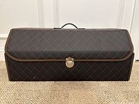 Closed view of rectangular trunk organizer showing black faux leather with red stitching in diamond quilted pattern, brown trim, metal clasp, and handle on top.