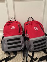 Photo of two red and grey backpacks with 72 HRS survival kit label, showing front and side profiles.