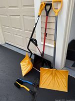 Photo of 3 snow shovels and 2 ice scraper brushes leaning against a garage wall on pavement.