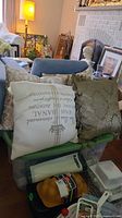 Four oversized decorative pillows arranged on and around a chair and storage bins. Includes cream pillow with gold crown and text and gold textured pillow.