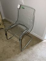 Side and angled view of a transparent acrylic chair with a chrome tubular metal base on carpeted floor.