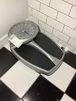 Oblique side angle photo of the grey and black large bathroom scale showing the circular dial with weight markings