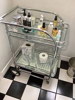 Full view of the chrome metal cart featuring two glass shelves, various bottles and containers on the top shelf for scale only, and a wire basket with a roll of toilet paper on the bottom shelf. Cart is placed on a black and white tiled floor next to a wall.