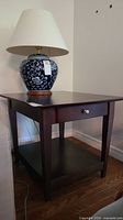 Solid wood end table shown with a ceramic lamp on top. Table has a dark finish, drawer with knob, and lower shelf. Visible light scratches on surface.