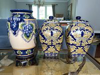 Front view of the ceramic grapevine vase with grapes in blue and green glaze, next to the pair of blue and gold floral ginger jars with lids.
