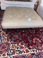 Top front angle view of the beige upholstered ottoman on a red patterned rug, showing full tufted surface and wood legs.