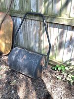 Full view of the metal lawn roller leaning against a wooden fence outdoors, showing drum and handle with aged wear and rust spots.