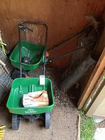 Wide view of two Scotts Turf Builder lawn seeders with green trays, black handles, and wheels, stored inside a wooden shed.