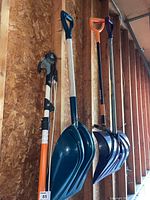 Four long-handled garden and winter tools hanging on a wall in a garage: Fiskars branch limber, two blue shuttle-shaped snow shovels with wooden handles, and a weed puller.