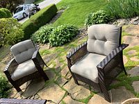 Two dark brown plastic wicker outdoor chairs side by side on stone patio with light grey seat and back cushions