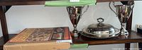 Full view of shelf showing boxed square pizza stone, silver plated covered serving tray, serving tray, and two large trophy style cups