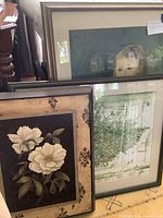 Photo showing three framed prints stacked against wall/based on floor; visible are the white flower print prominent in the front, large print with child portrait behind, and map print partially visible.
