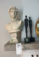Bust of David resin sculpture with pedestal, next to two tall slender black statues on square bases, partially showing a gold-framed mirror.