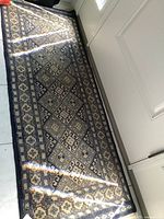 Top view of the full length of the runner rug on tile floor next to a door, showing the intricate geometric and floral repeating pattern in blue and beige with fringed ends.