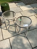Pair of beige metal framed round patio side tables with clear textured glass tops pictured on stone patio tiles outdoors.