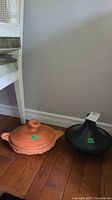 Two tangines on hardwood floor next to white chair. One tangine is natural clay color, the other is black stoneware.