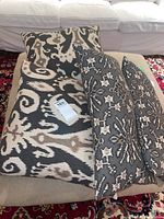Photo showing folded throw pillows stacked on a beige ottoman; includes two larger square pillows and two smaller rectangular pillows with black, beige, and cream designs.