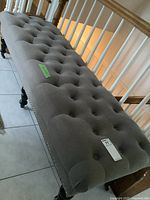 Side angled view of grey upholstered tufted hall bench showing overall length, seat tufting, nail head trim, and wood legs with casters.