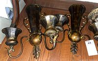 Photo showing two dark metallic wall sconces with three holders each, ornate scrollwork arms, and decorative motifs on wooden floor.