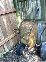 Full view of older metal wheelbarrow leaning against a fence showing metal tub and soft tire.
