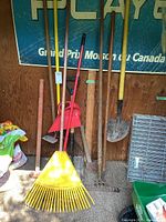 Full view of yard tools arranged against wall including rakes, shovels, axe, broom, and other tools