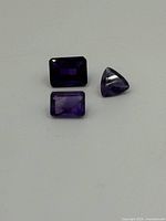 Three loose amethyst gemstones placed flat on a white surface, showing faceted tops and different shapes.