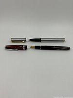 Top view of Shaefer ballpoint pen with silver-tone barrel and cap with gold accents alongside the Modele Recife Depose marbled burgundy fountain pen with silver and gold nib.